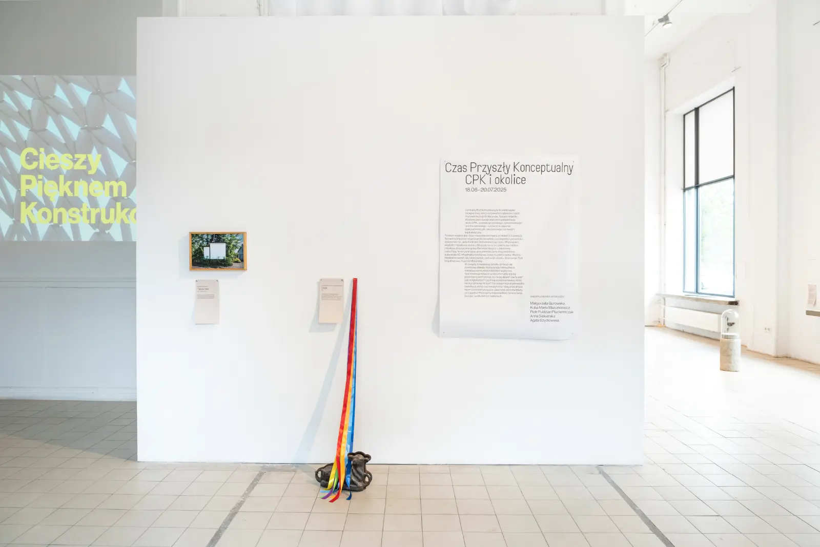 Widok wystawy „Czas Przyszły Konceptualny. CPK i okolice”. Na białej ścianie widoczny jest tekst kuratorski, instalacja z kolorowymi wstążkami i fotografia w drewnianej ramie.