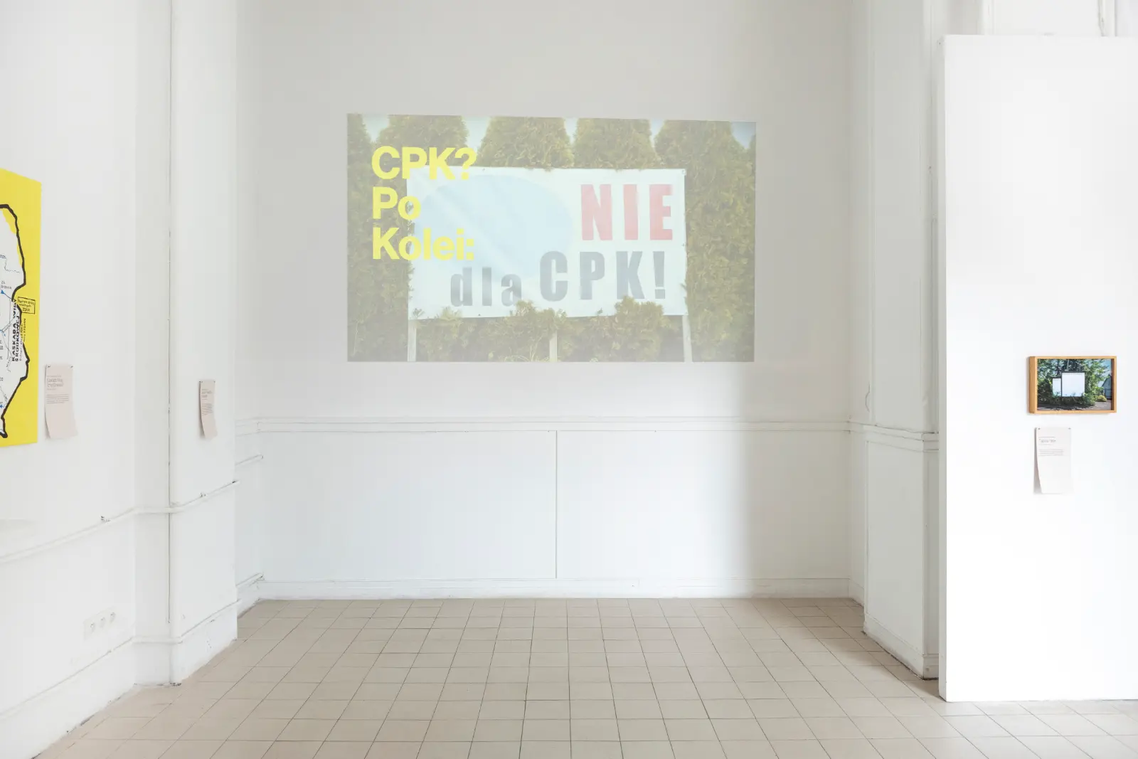 Kadr z pracy wideo wyświetlony na białej ścianie galeryjnej. Po lewej stronie widać fragment żółtego obrazu, po prawej fotografię w drewnianej ramie.