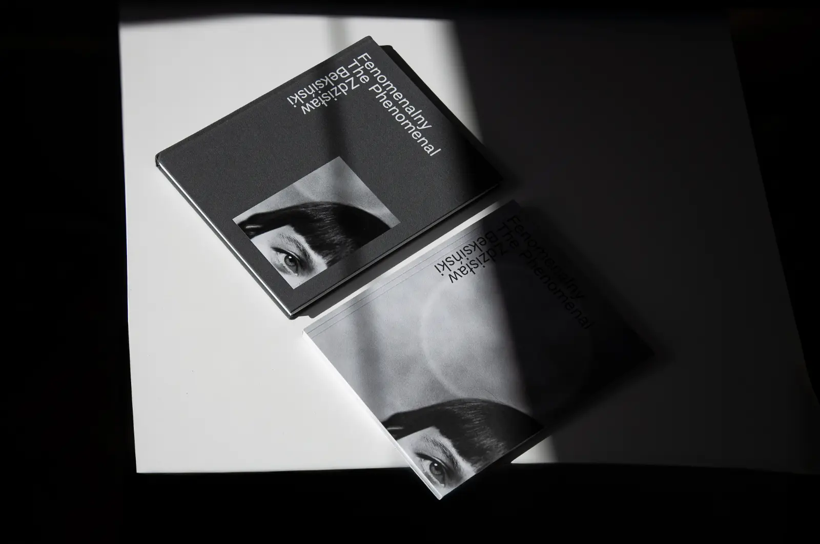 Dwie książki na białym blacie. Jedna z nich jest częściowo w cieiu. Na obu znajduje się minimalistyczna typografia i portret kobiety – żony Beksińskiego.