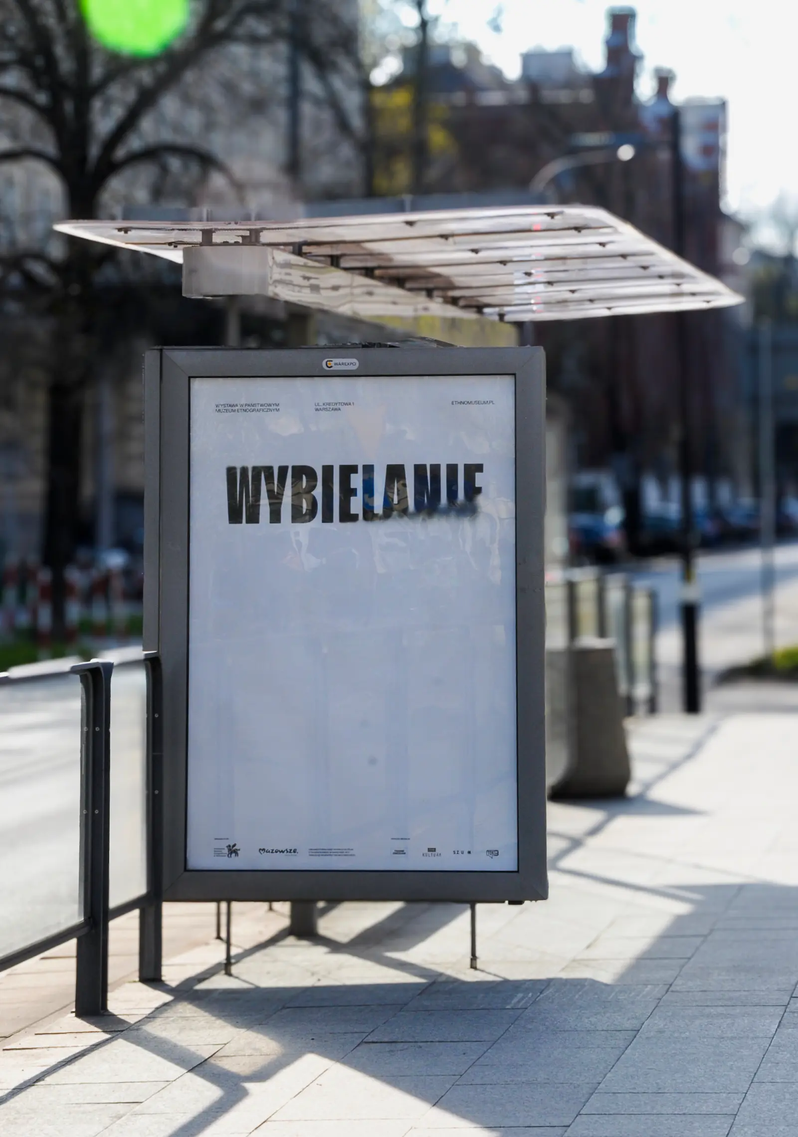 A poster for the “Bleaching” exhibition at a tram stop on a sunny day on a Warsaw street.